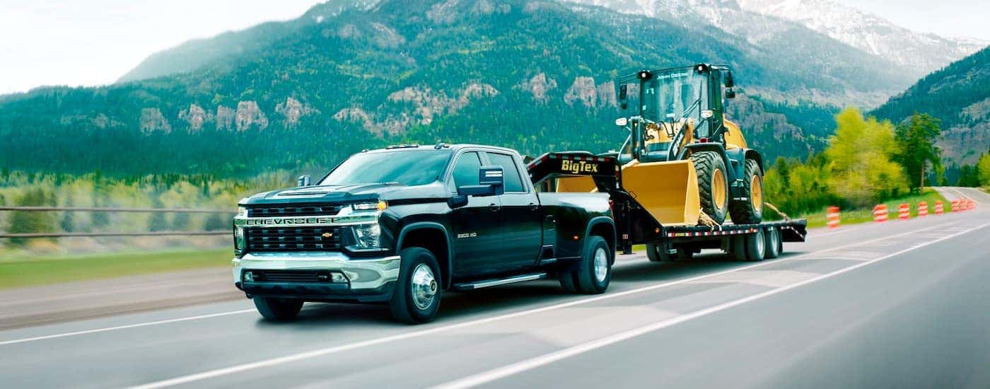 A black 2021 Chevy Silverado 3500 HD is shown from the side towing a trailer with construction equipment down an empty highway.