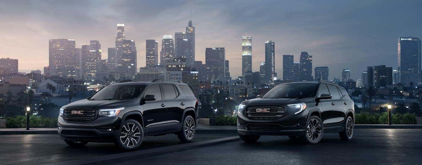 Two 2019 GMC Black Edition SUVs, a Terrain and an Acadia, are shown at night with a city in the distance.