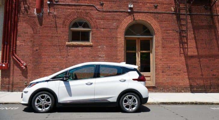 A popular used car for sale near you, a white 2020 Chevy Bolt EV, is parked on the street next to a brick wall.