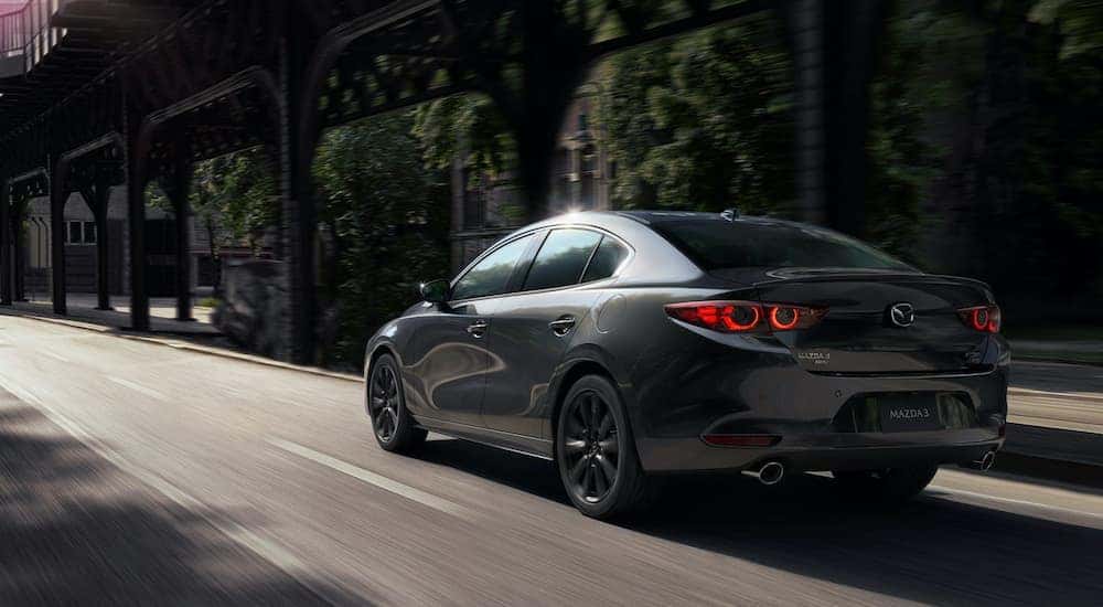A popular used car for sale near you, a gray 2020 Mazda 3 sedan, is shown from the rear driving down a city street.