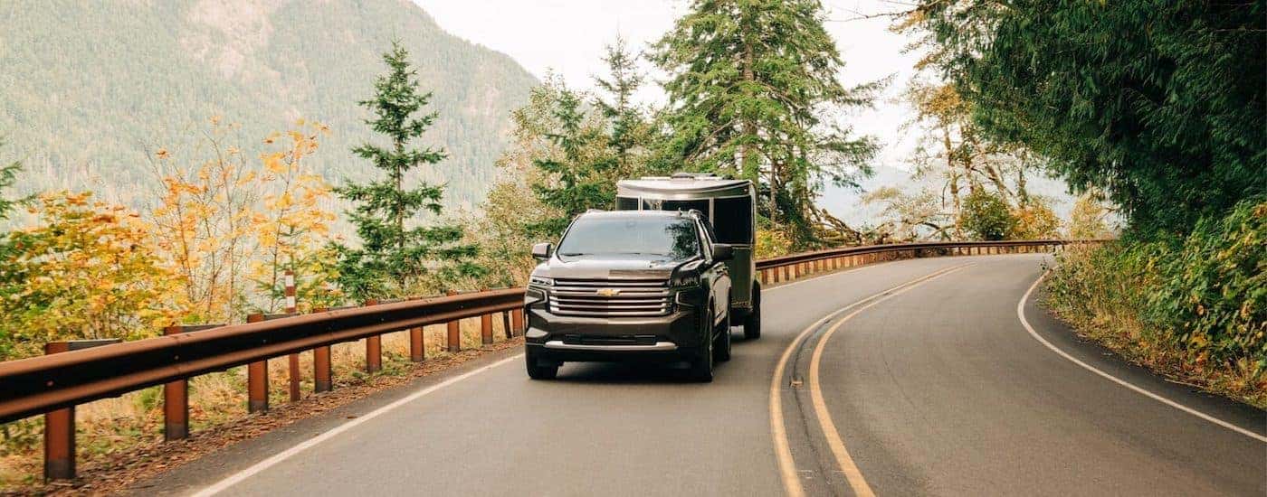 A black 2021 Chevy Tahoe High Country is shown towing a trailer down a wooded road in the mountains.