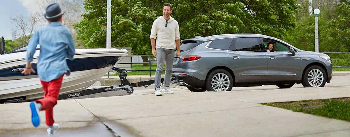 A grey 2022 Buick Enclave is shown towing a boat near a child running.