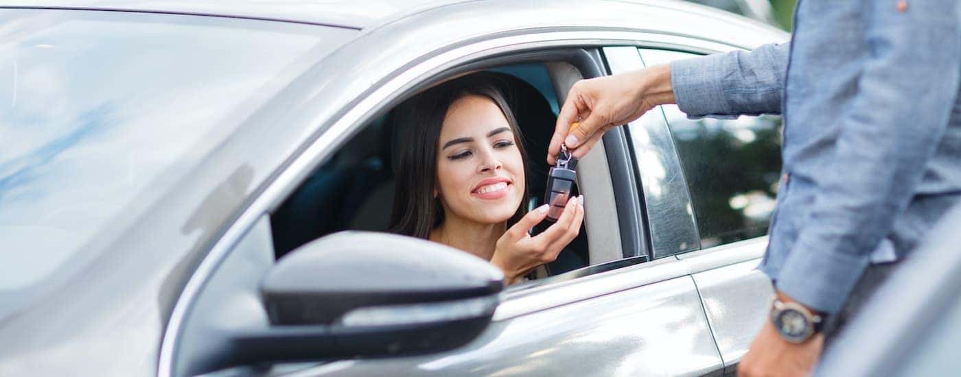 A salesperson is shown passing a car key to a woman in the drivers seat of a car.