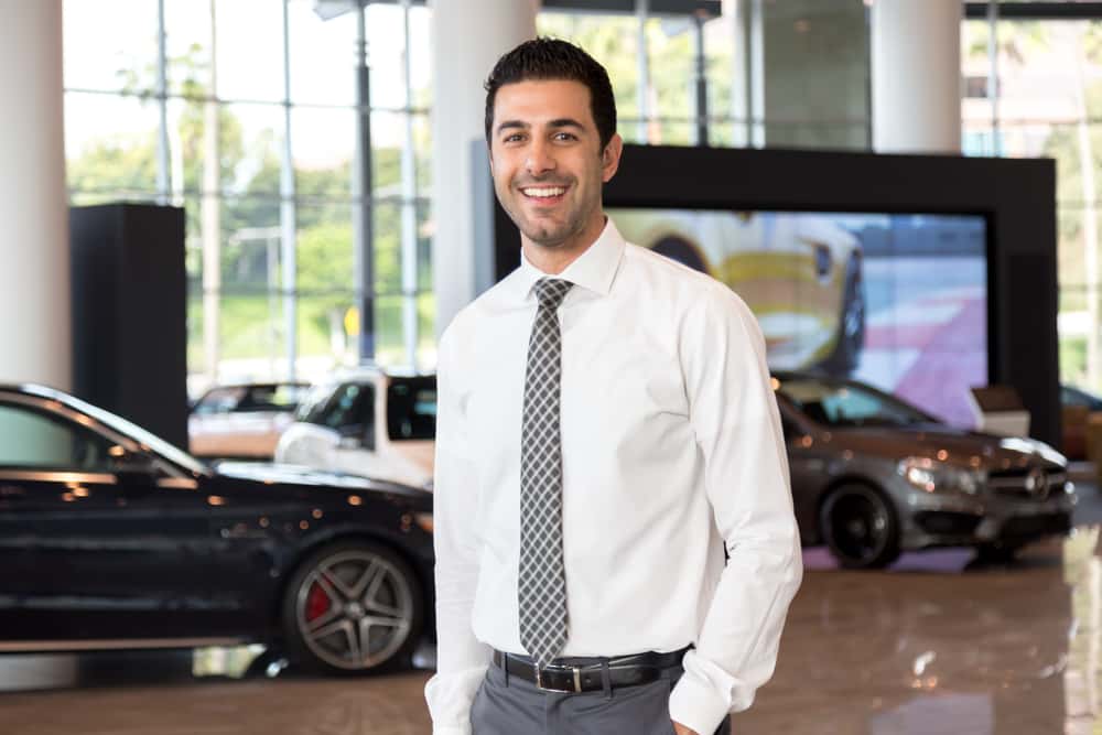 A tall man smiling and standing in the dealership with a white shirt and a checkered tie