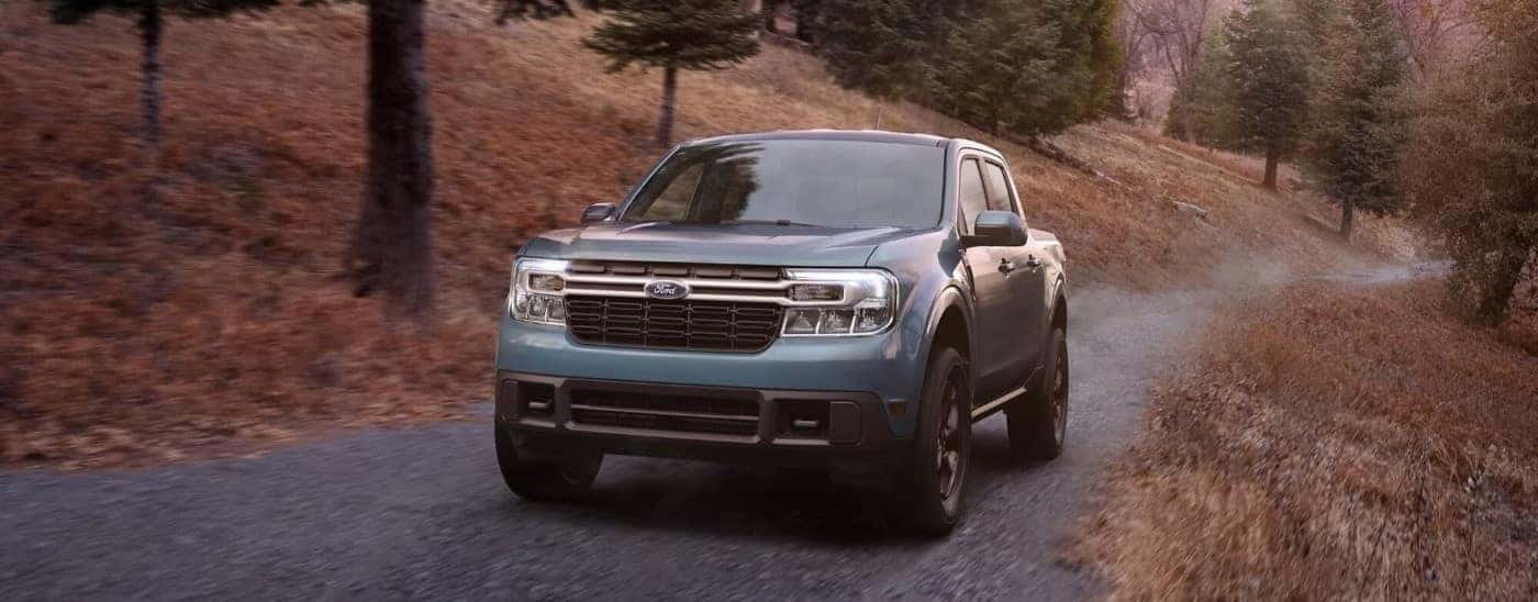 A blue 2023 Ford Maverick driving up a gravel road in the forest