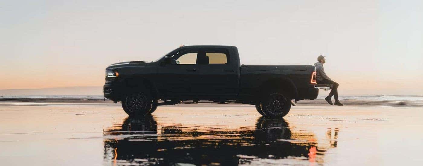 A man sitting on the bed of a black 2023 Ram 2500 parked on a beach