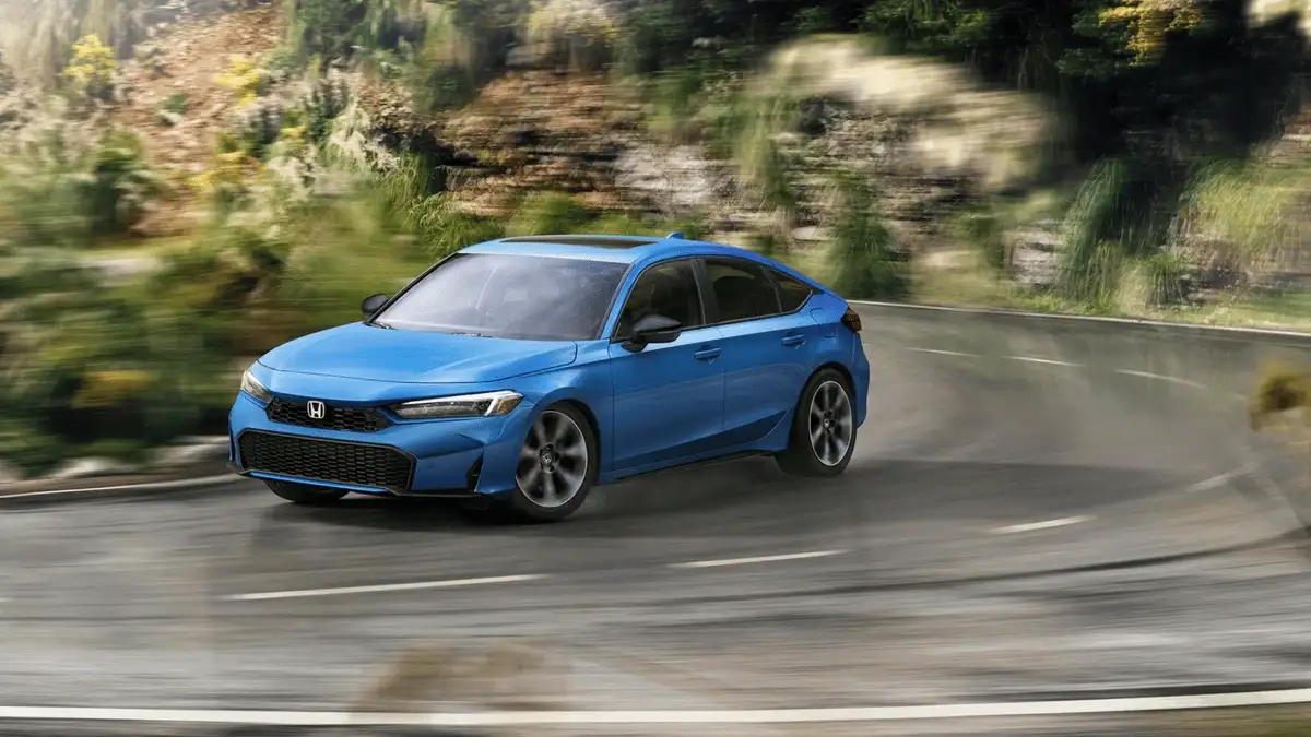 2026 Honda Civic Hatchback Hybrid near Smithtown, NY blue hatchback taking a curve on a scenic road, front three-quarter view