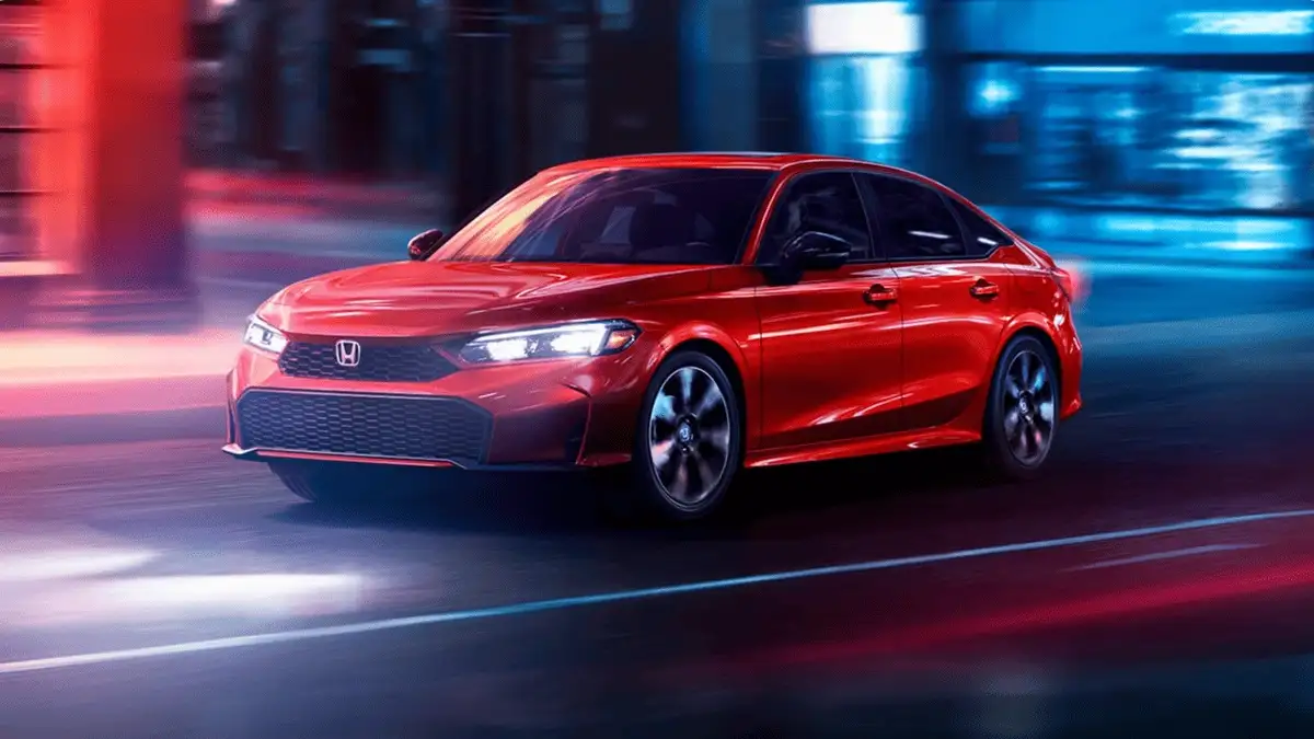 2026 Honda Civic Sedan Hybrid near Smithtown, NY red sedan driving through city lights at night, front three-quarter view