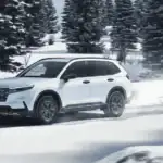 2026 Honda CR-V Hybrid in Platinum White Pearl driving through snowy road — fuel economy faceoff for long drives near Commack NY vs Ford Escape Hybrid