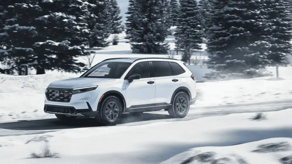 2026 Honda CR-V Hybrid in Platinum White Pearl driving through snowy road — fuel economy faceoff for long drives near Commack NY vs Ford Escape Hybrid