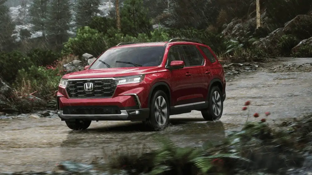 2025 Honda Pilot in Radiant Red Metallic front three-quarter view driving through rainy forest trail near Commack NY