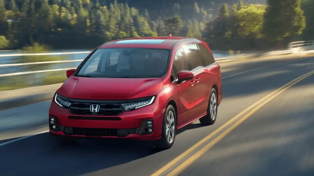 2026 Honda Odyssey minivan in Radiant Red Metallic front three-quarter view driving on scenic forest highway