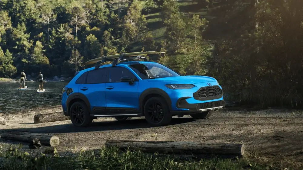 Blue 2026 Honda HR-V with a surfboard on the roof rack parked by a lakeside trail, a great first SUV near Hauppauge NY