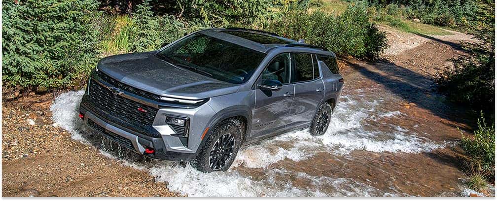Gray 2026 Chevrolet Traverse driving through a stream, viewed from a front three-quarter angle.