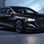 A black 2021 Chevy Malibu is shown parked in front of a modern building after leaving a Chevy dealer near you.