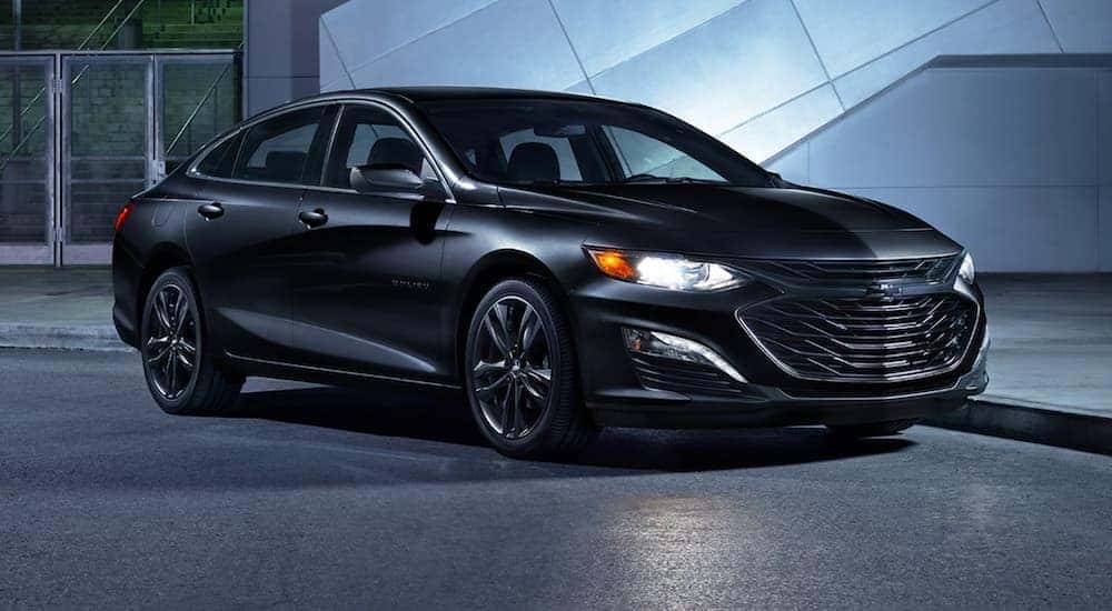 A black 2021 Chevy Malibu is shown parked in front of a modern building after leaving a Chevy dealer near you.
