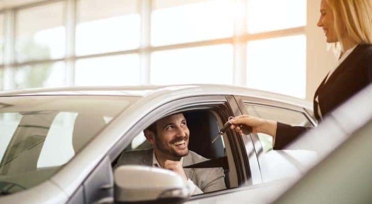 A close up shows a customer in a car being handed car keys by a saleswoman.