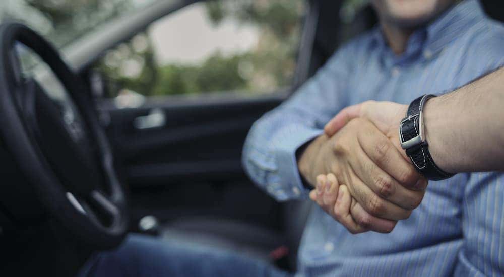 A person is sitting in the drivers seat of a car shaking hands with a salesperson.