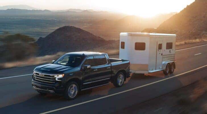 A black 2023 Chevy Silverado 1500 High Country is shown towing a white trailer on an open highway.