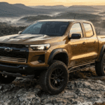 Gold Chevrolet Colorado ZR2 pickup truck parked on a rocky cliff overlooking a misty mountain valley at sunrise.