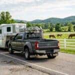 A realistic wide photograph of a heavy-duty crew-cab pickup truck hitched to a three-horse gooseneck trailer on a rural Tennessee two-lane road, morning light, white board fencing and a green pasture with grazing horses along the roadside, gooseneck hitch and trailer coupling visible in clear detail, low rolling hills behind, documentary photographic style. No text, no logos other than natural vehicle badging, no identifiable faces. (Representative image for illustration purposes only. Actual vehicle may vary based on trim and configuration.)