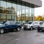 A realistic wide photograph of freshly delivered new vehicles arriving at a dealership, a full-size pickup and two crossover SUVs staged on clean pavement in front of a modern glass showroom facade, protective transit film still on a hood edge, bright overcast daylight, autumn trees reflected in the glass, crisp commercial automotive photography. No text, no signage lettering, no logos other than natural vehicle badging, no identifiable faces. (Representative image for illustration purposes only. Actual vehicle may vary based on trim and configuration.)