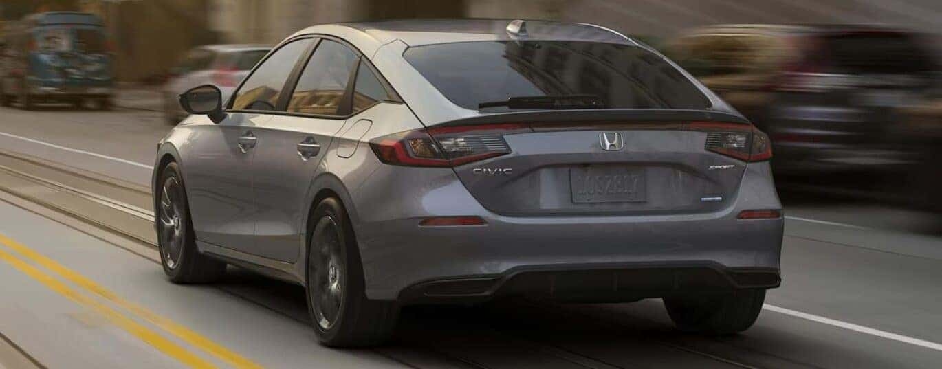A gray 2025 Honda Civic Hatchback Sport Touring Hybrid is shown driving in a city.