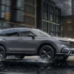 A grey 2025 Honda CR-V Sport Touring Hybrid is driving on a city street in the rain.