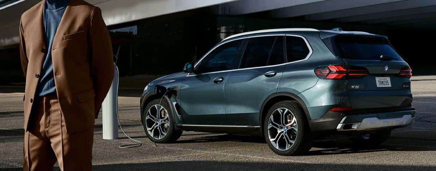 A blue 2025 BMW X5 is charging on the side of a city street.