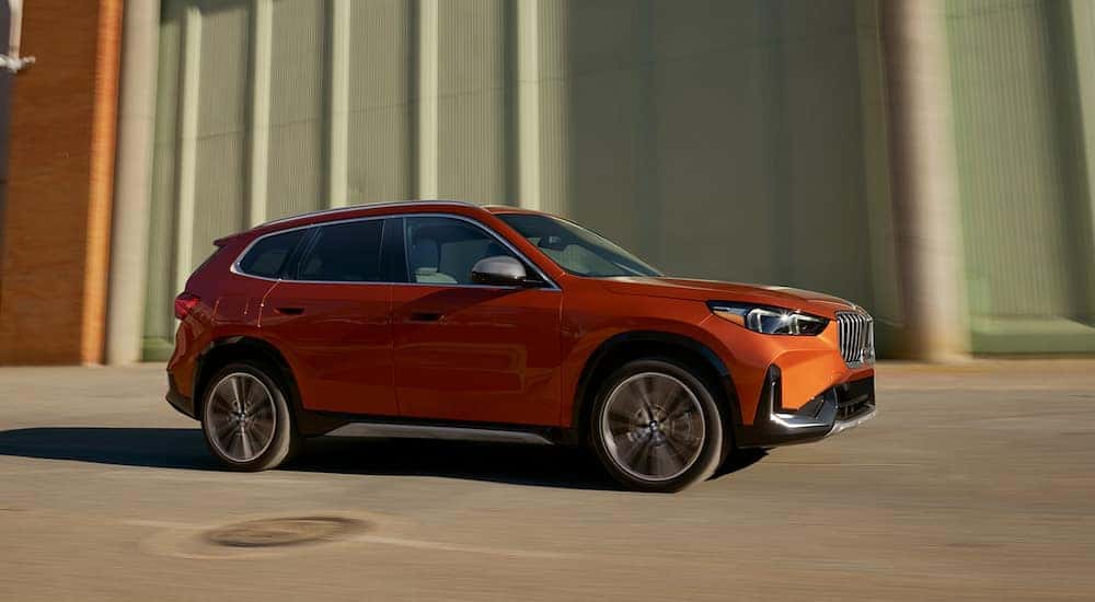 Side view of an orange 2023 BMW X1 driving past buildings.