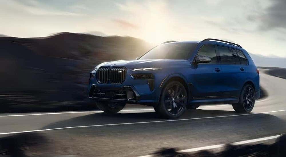 A blue 2023 BMW X7 M60i is shown driving on a winding road.