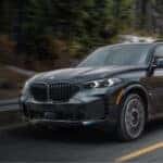 A grey 2025 BMW X5 for sale is shown driving on a tree-lined road.