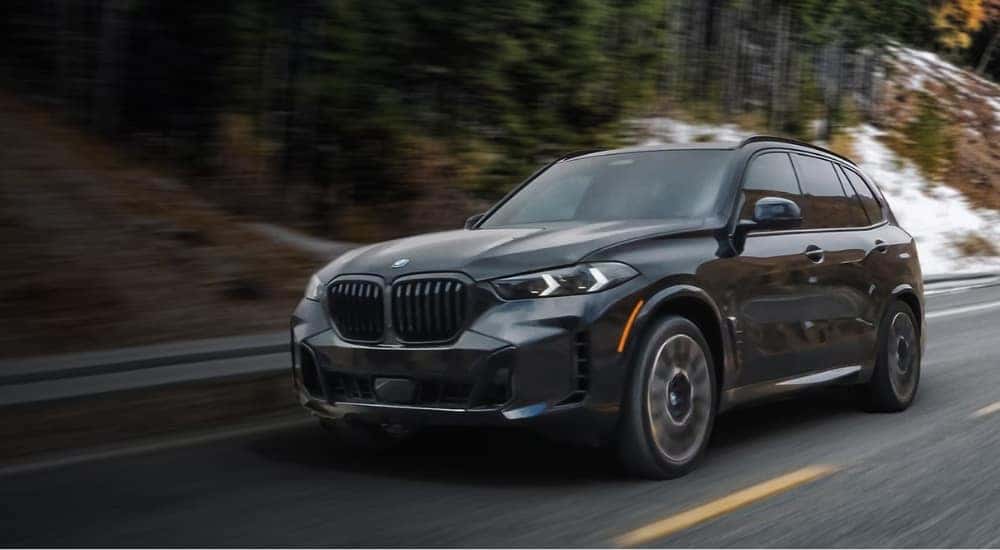 A grey 2025 BMW X5 for sale is shown driving on a tree-lined road.