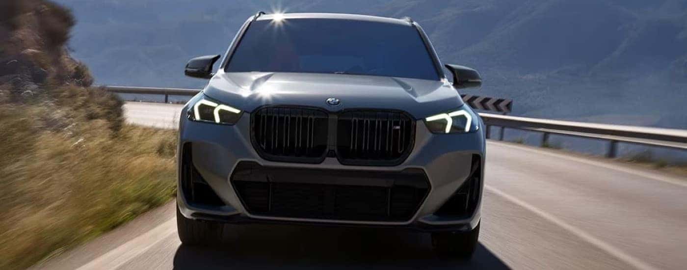 A grey 2024 BMW X1 is shown from the front driving on a mountain road.