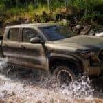 A bronze 2024 Toyota Tacoma from the side after leaving a dealer that has a lifted truck for sale.