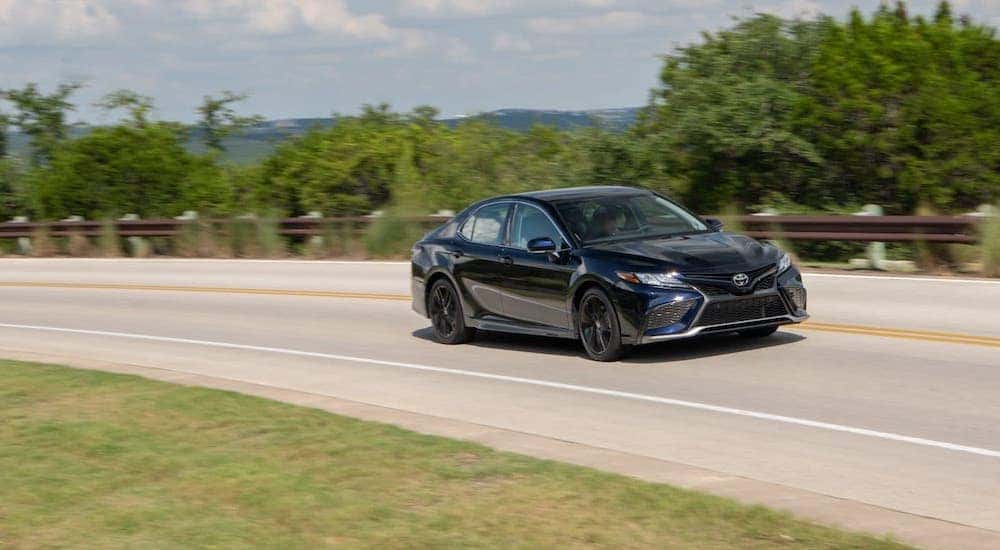 A black 2021 Toyota Camry is driving on a highway in front of trees after leaving a Toyota Camry dealer.