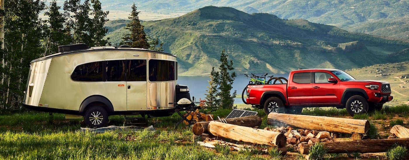 A red 2021 Toyota Tacoma is parked at a campsite with a small, metal camper in front of a lake and mountains.
