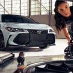 Two mechanics are shown checking the suspension on a white 2021 Toyota Avalon TRD.