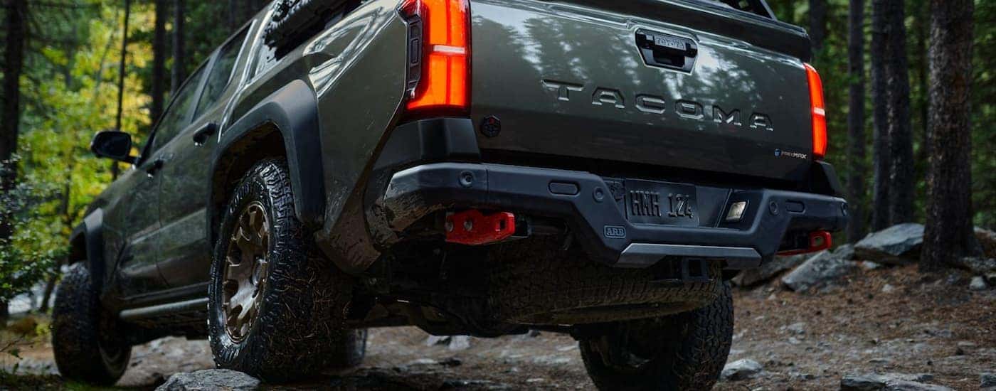 A green 2024 Toyota Tacoma Trailhunter is shown from the rear driving on a forest trail.