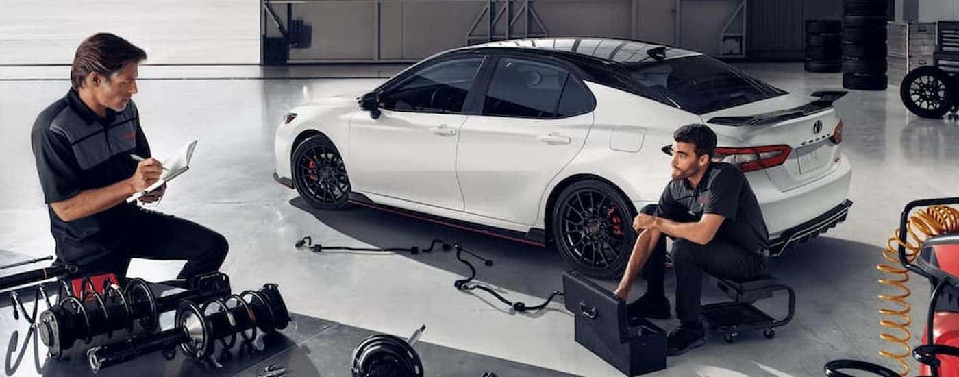 Two mechanics are shown upgrading parts on a white 2022 Toyota Camry TRD.