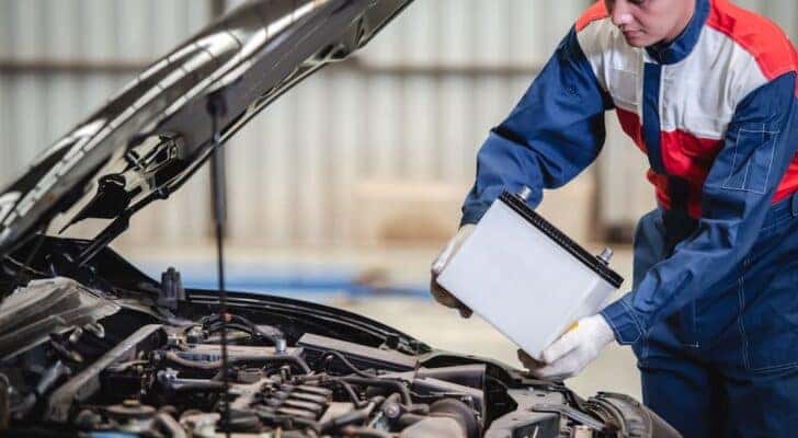 A mechanic is shown holding a car battery during a battery replacement.