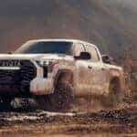 One of many popular Toyota trucks for sale, a white 2024 Toyota Tundra, is shown driving on mud.