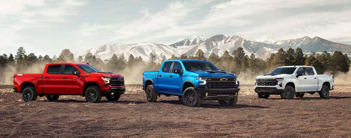 A red, a blue, and a white 2024 Chevy Silverado 1500 are parked on a dirt lot.