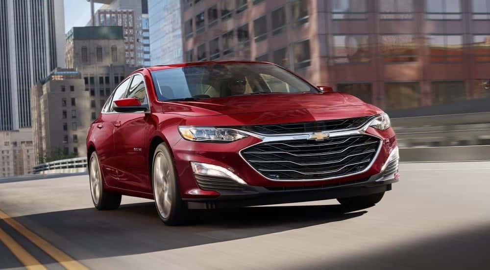 Front angle view of a red 2025 Chevy Malibu driving through a city.