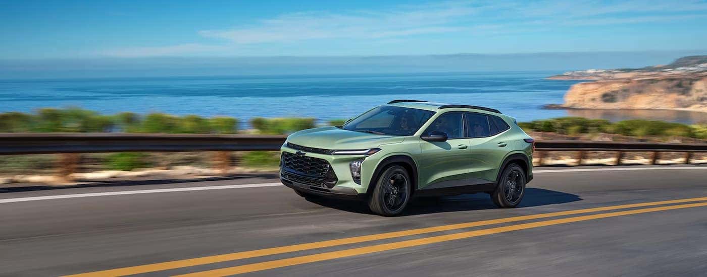 A green 2025 Chevy Trax driving down a highway near a lake