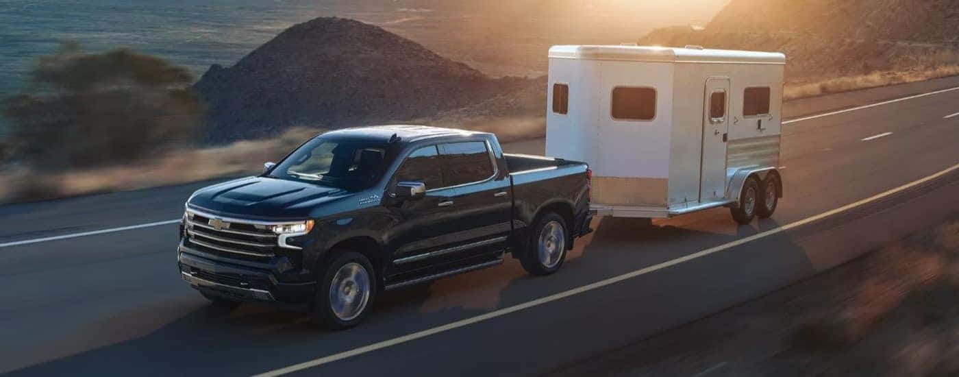 A black 2023 Chevy Silverado 1500 High Country is towing a white horse trailer on a mountain road.
