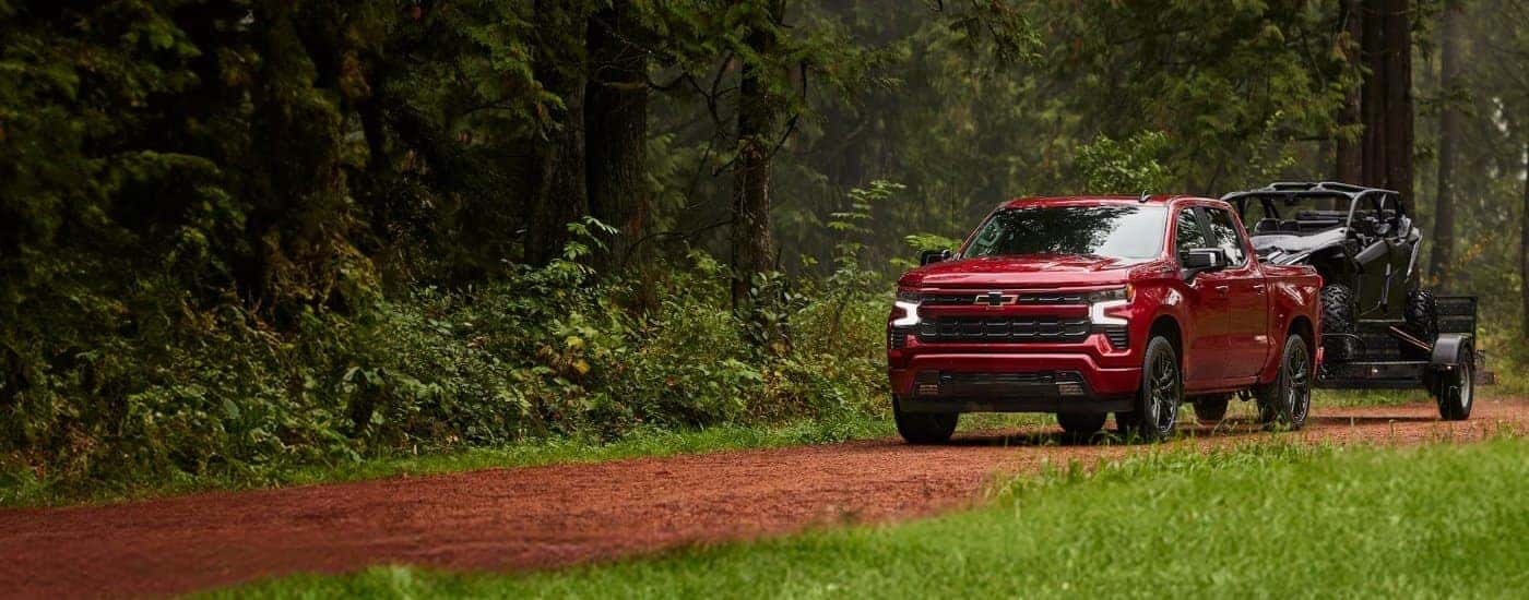 A red 2025 Chevy Silverado 1500 RST is shown towing a trailer off-road.