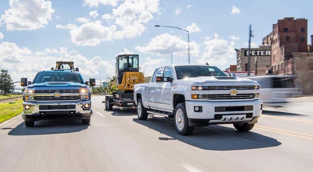 Two 2018 Chevy Silverado 2500 HD towing heavy machinery down the road.