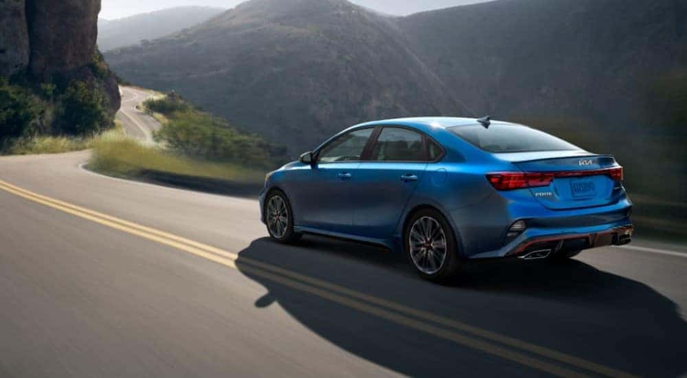 A blue 2023 Kia Forte is shown driving on a highway near mountains.