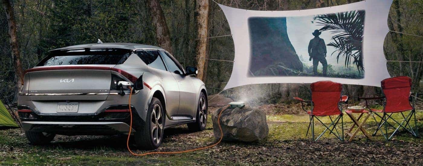 A silver 2024 Kia EV6 is shown parked at a campsite while a movie plays through a projector.